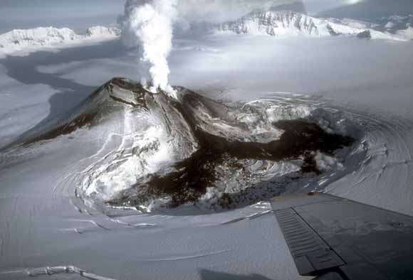 photo of volcano taken from a low-flying aircraft.  Steam is coming out of the top and snow covers the surrounding region.