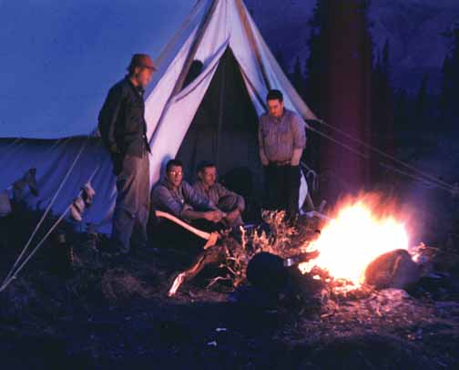 photo, four men in the evening light outside of a tent in front of a campfire