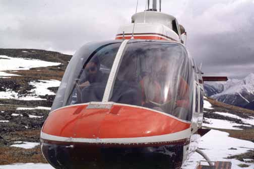 photo, Bill in the front seat of a parked helicopter.  Sparse snow cover on the ground