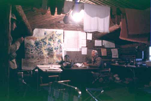 photo, Bill at a table in a cabin with maps on the wall and laundry hung from the ceiling