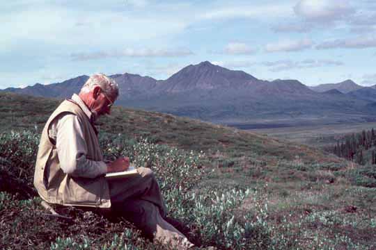 photo, Bill taking notes, sitting on a hillside with mountains in the background